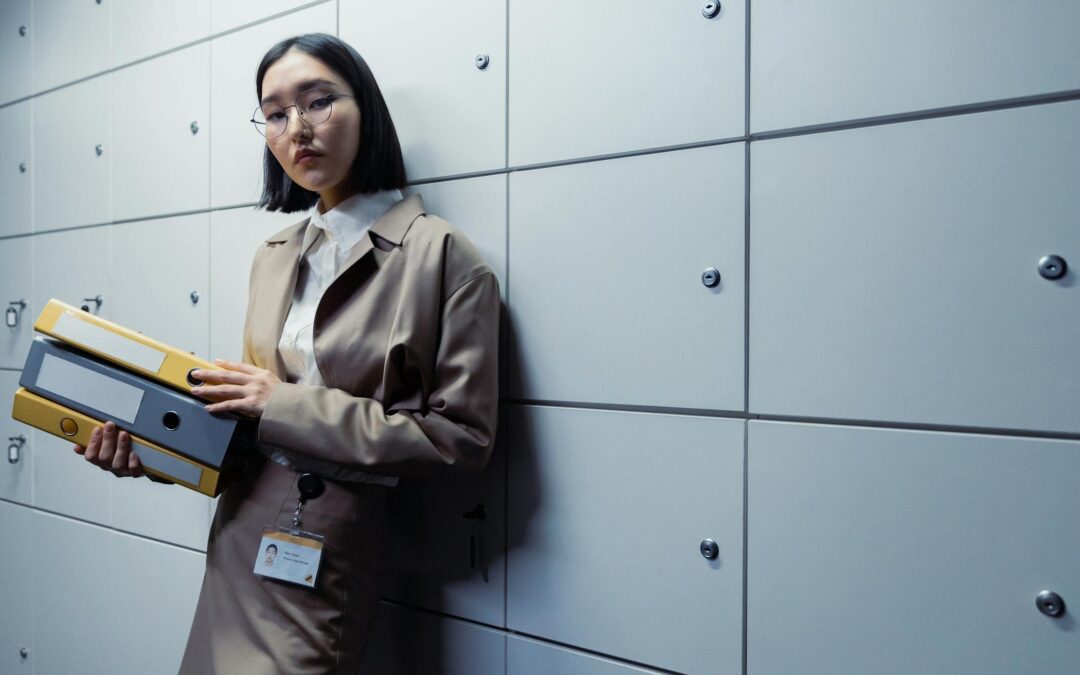 Image depicting a woman of ethnicity, Asian with glasses. Leaning on a wall of storage, while holding binders